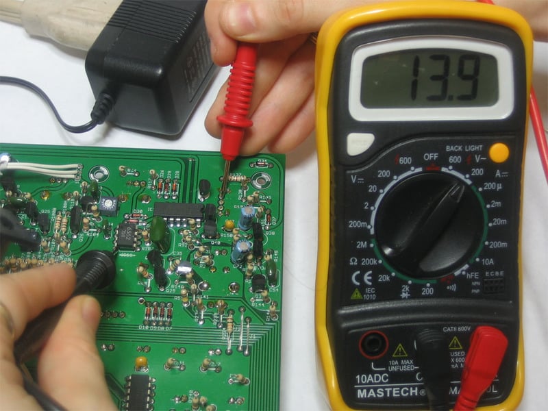 Multimeter measuring a powered circuit board, displaying 13.9.